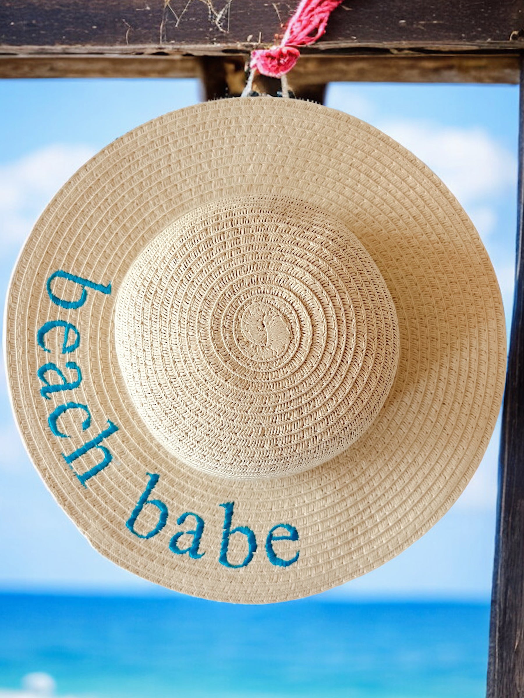 Woven straw hat with "beach babe" embroidered, hanging against a blue ocean backdrop.
