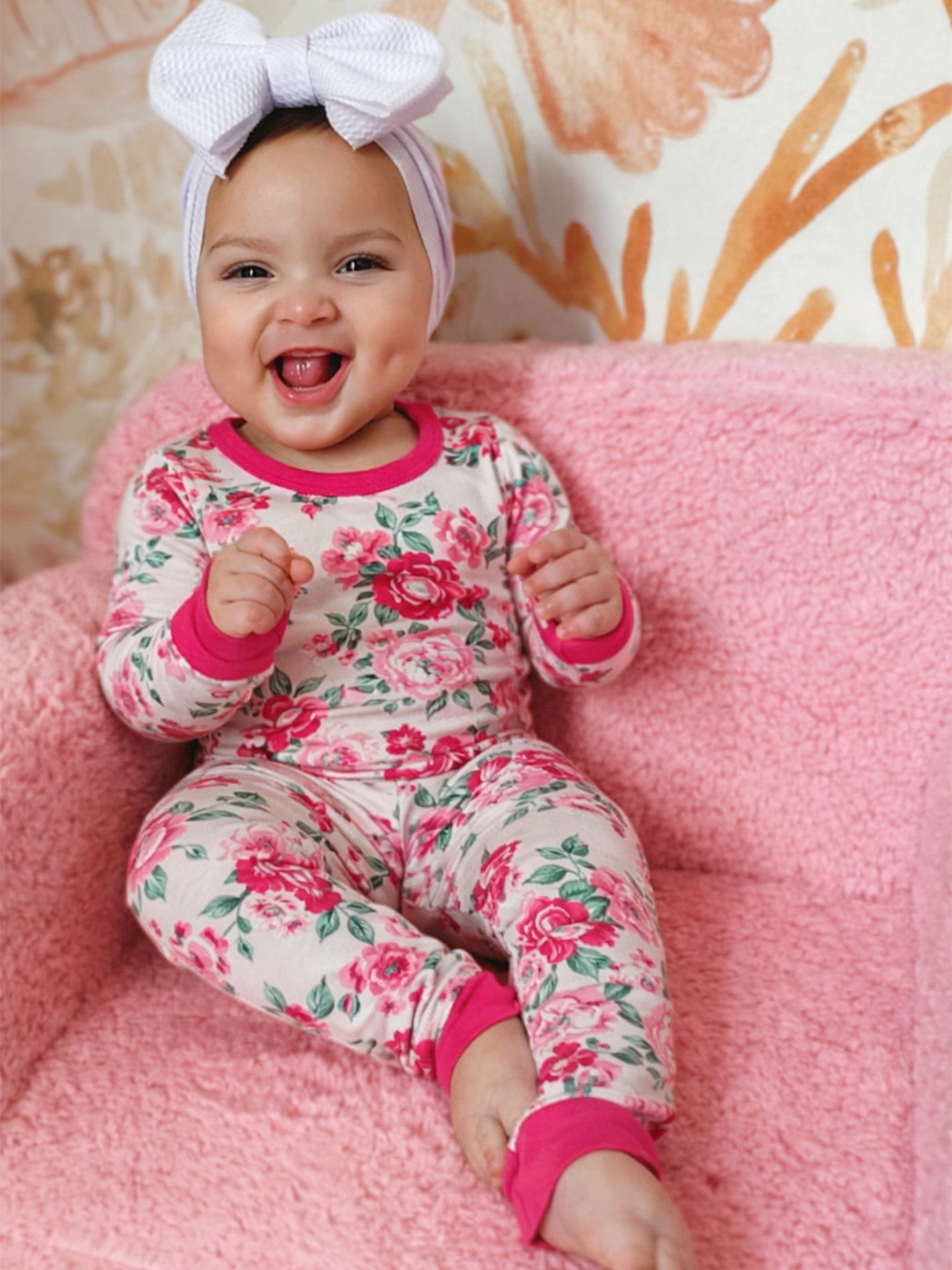 Smiling baby in floral pajamas sitting on a pink chair with a large white bow headband.