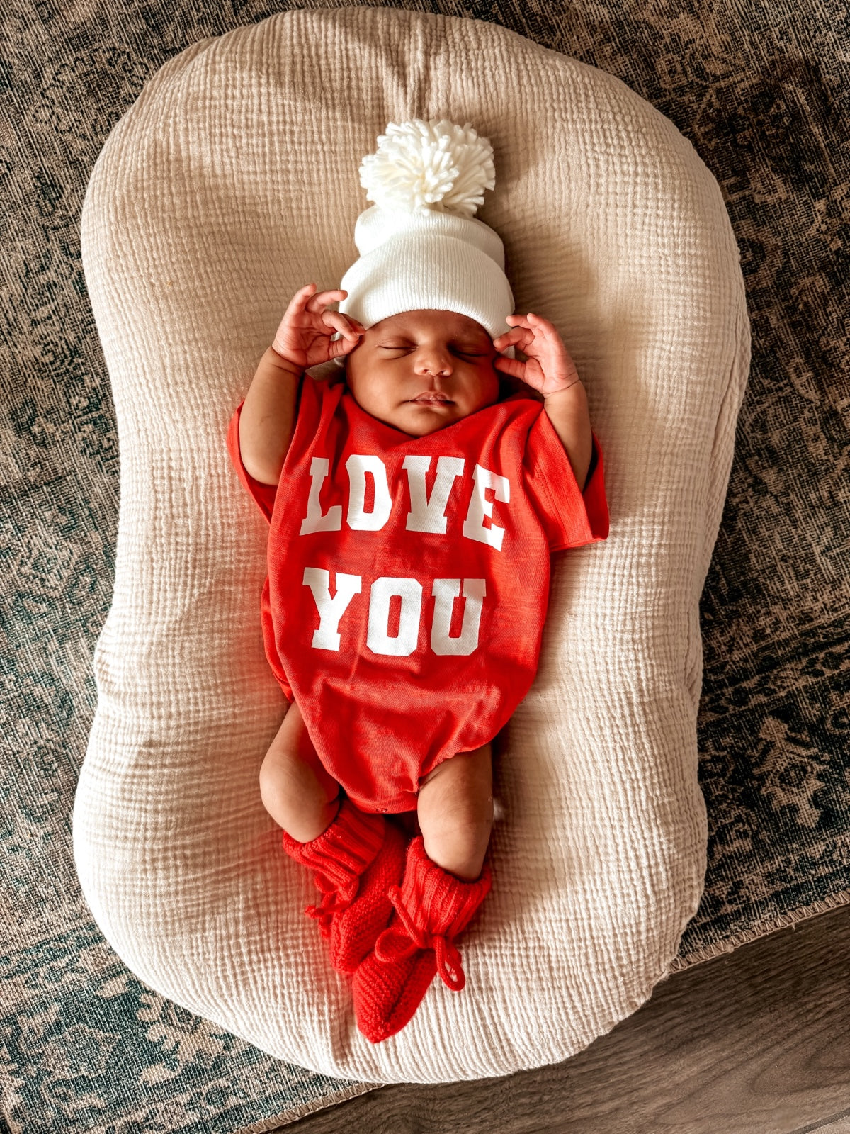 Infant lying on a cushioned surface, wearing a red "LOVE YOU" outfit, white hat, and red booties.