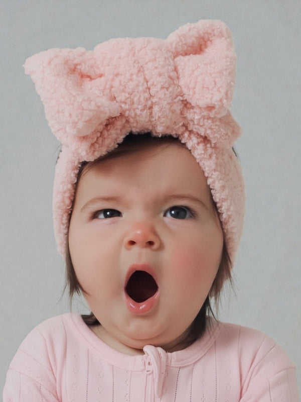 Baby girl in a pink headband yawning while wearing a pink outfit, with a neutral background.