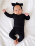Smiling baby in black outfit and bow headband, lying on a white bed with arms and legs outstretched.