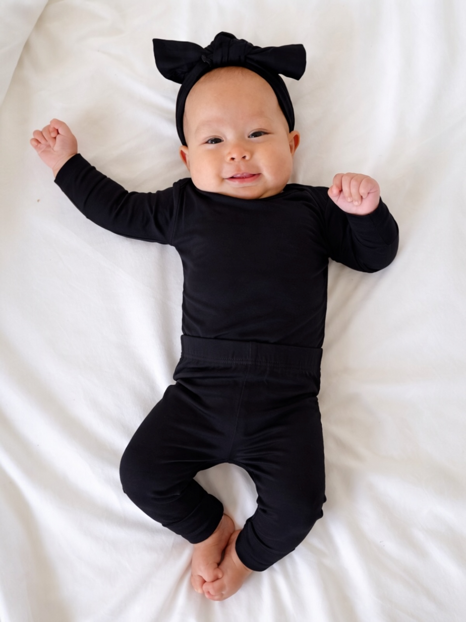 Smiling baby in black outfit and bow headband, lying on a white bed with arms and legs outstretched.
