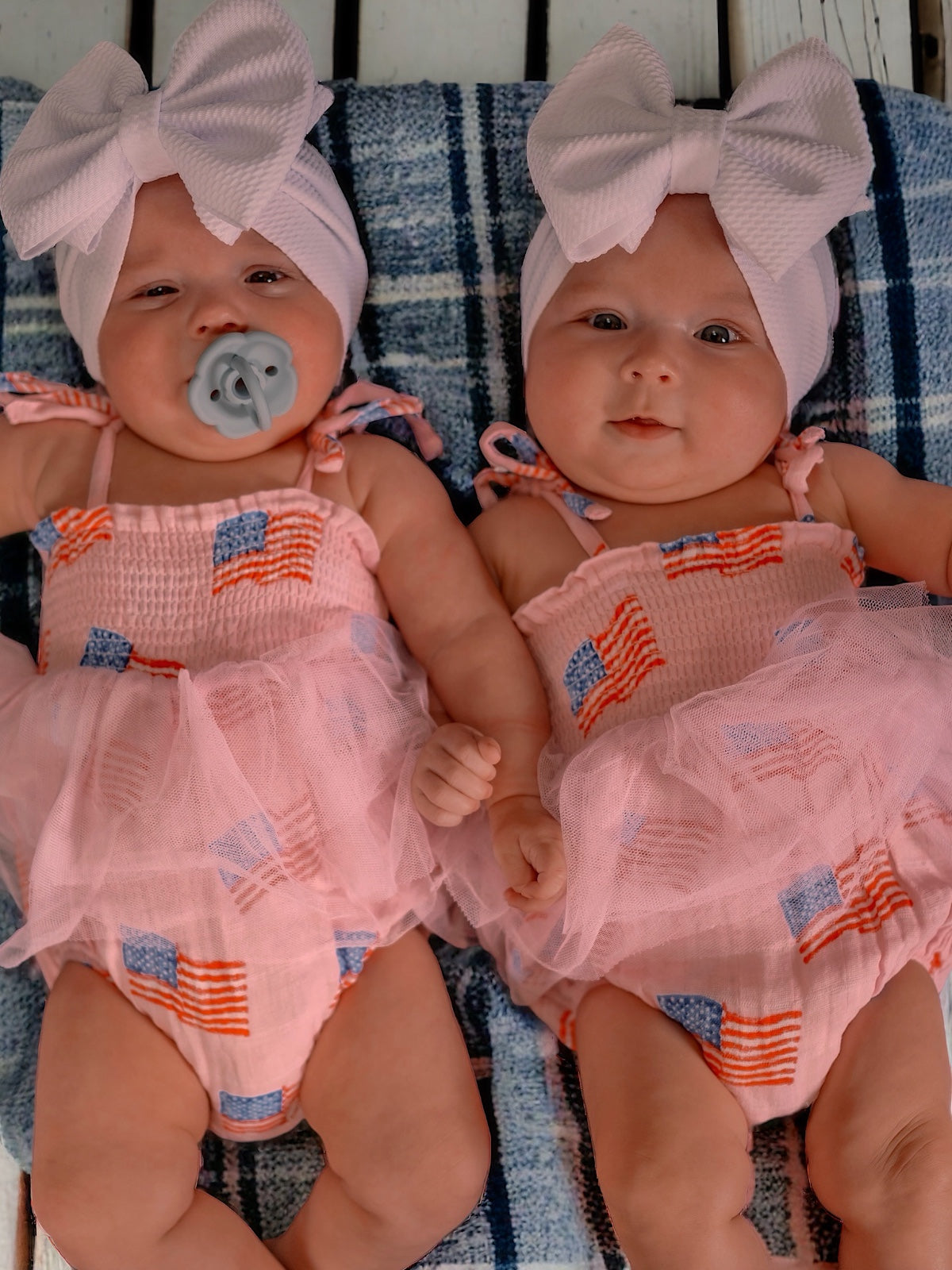 Twin babies wearing pink dresses with American flags, large bows, and pacifiers, lounging on a plaid blanket.