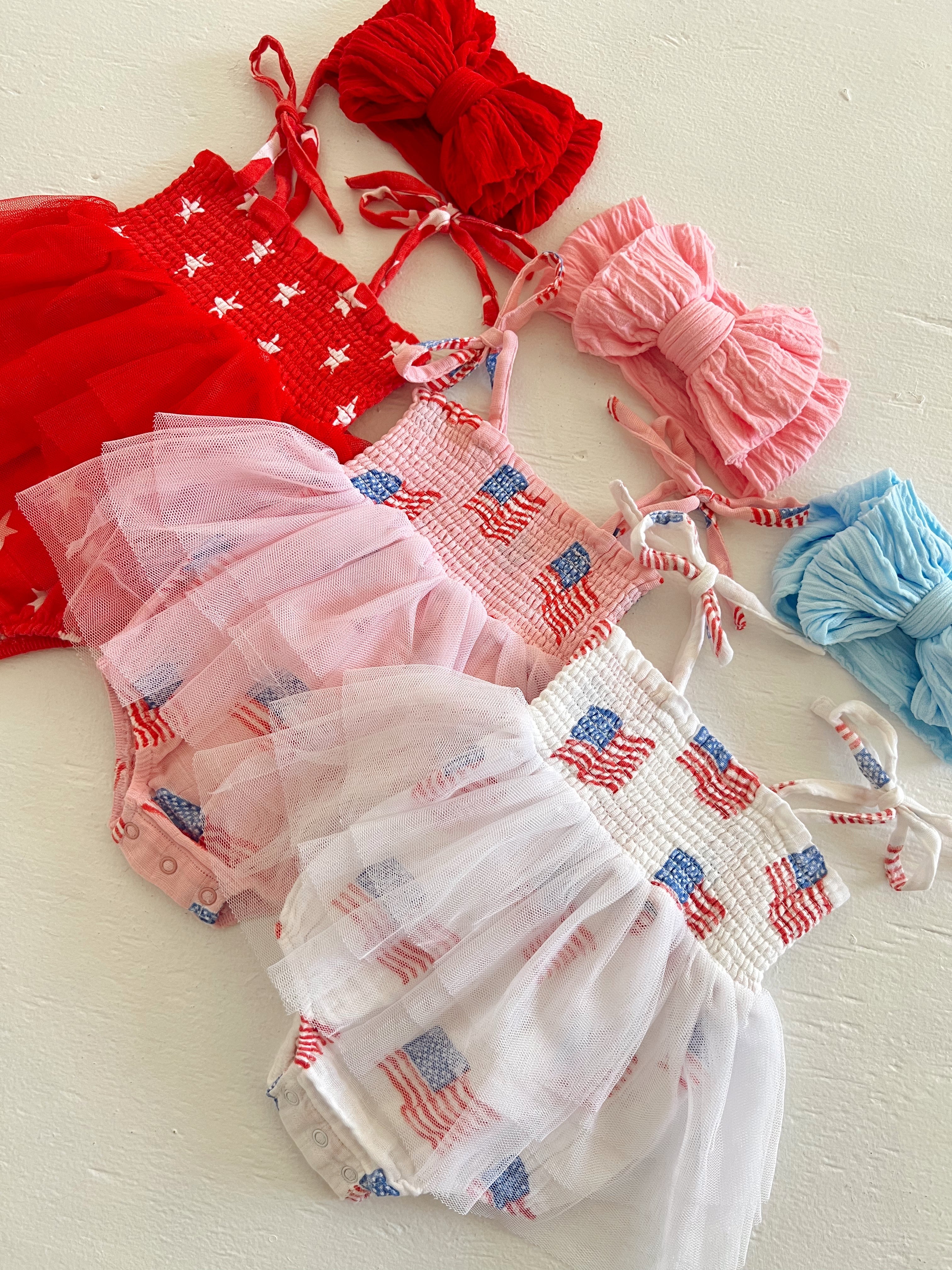 Colorful toddler outfits with red, white, and blue patterns, featuring ruffles and bows on a light background.