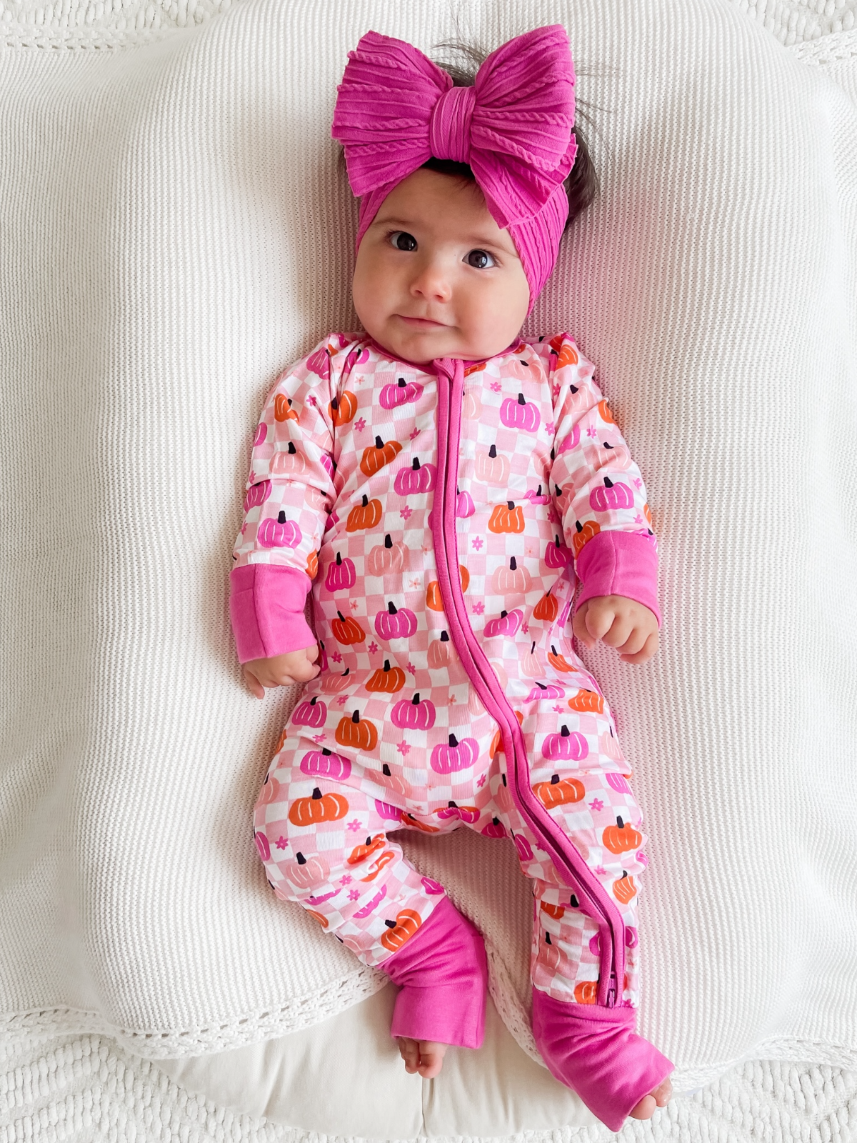 Baby in pink pumpkin-patterned onesie and large pink bow on a textured white blanket.