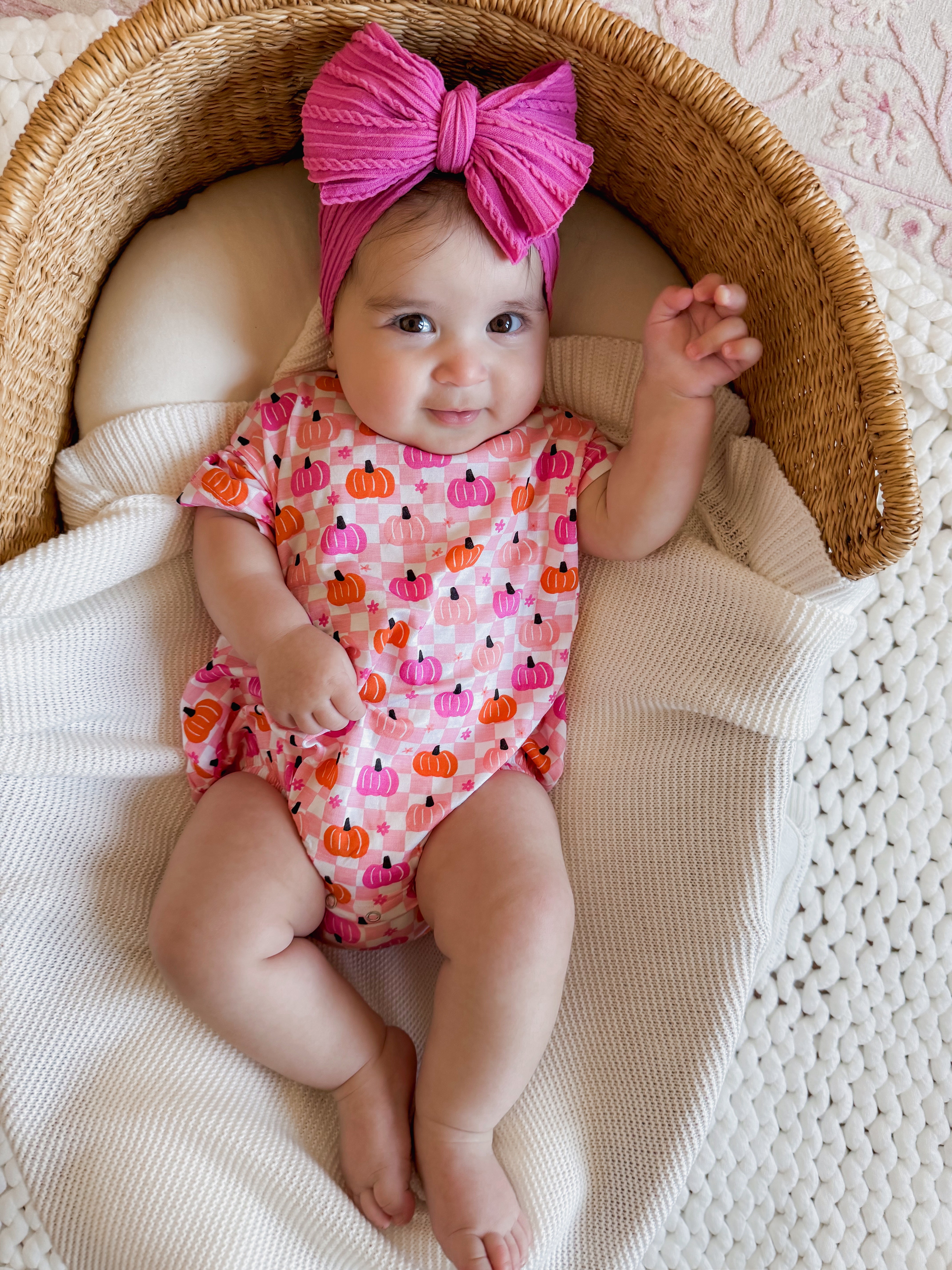 Smiling baby in a pumpkin-patterned outfit and pink bow, lying in a cozy basket on a textured blanket.