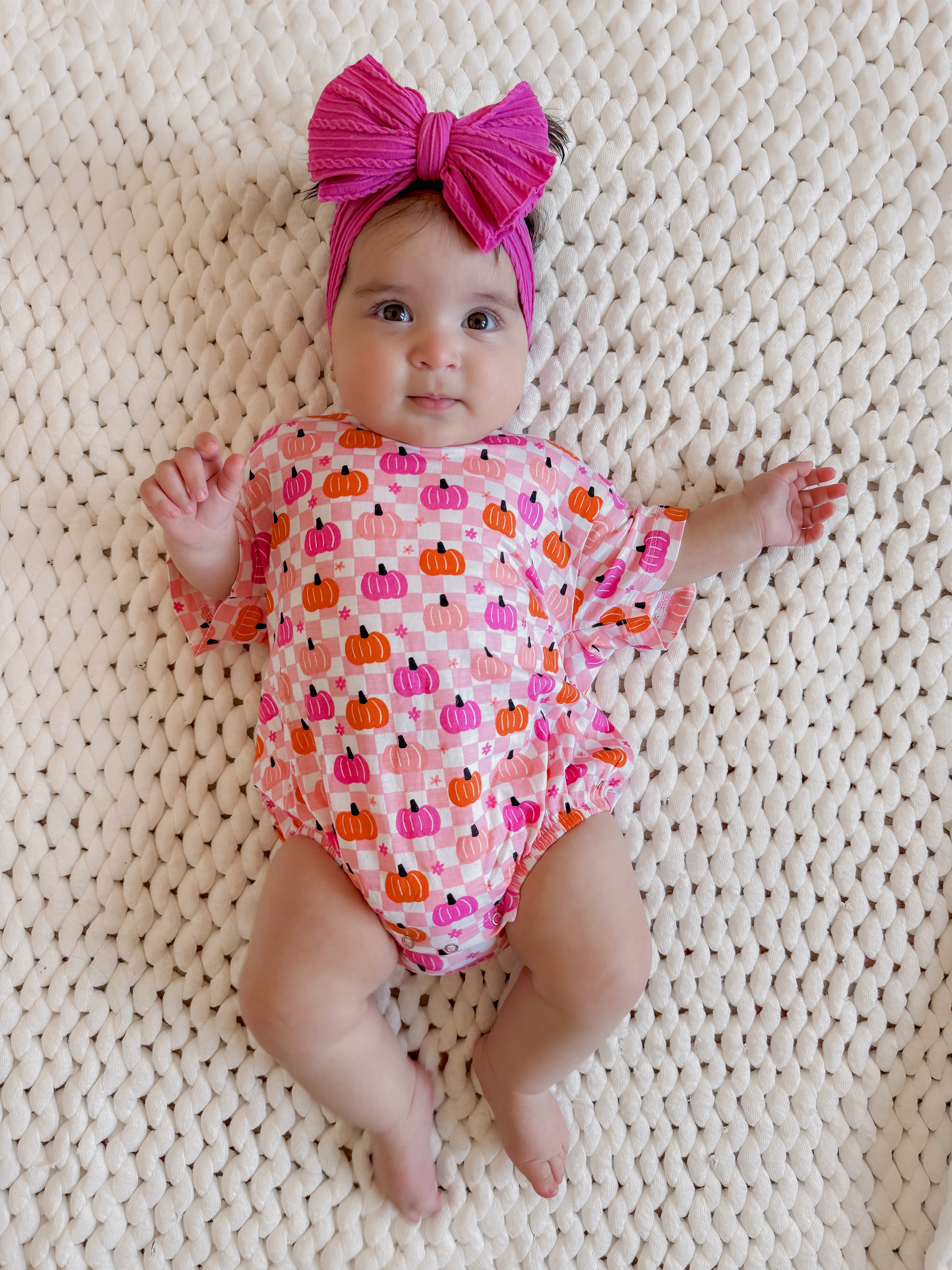 Baby in a pumpkin-patterned outfit with a large pink headband, lying on a textured blanket.