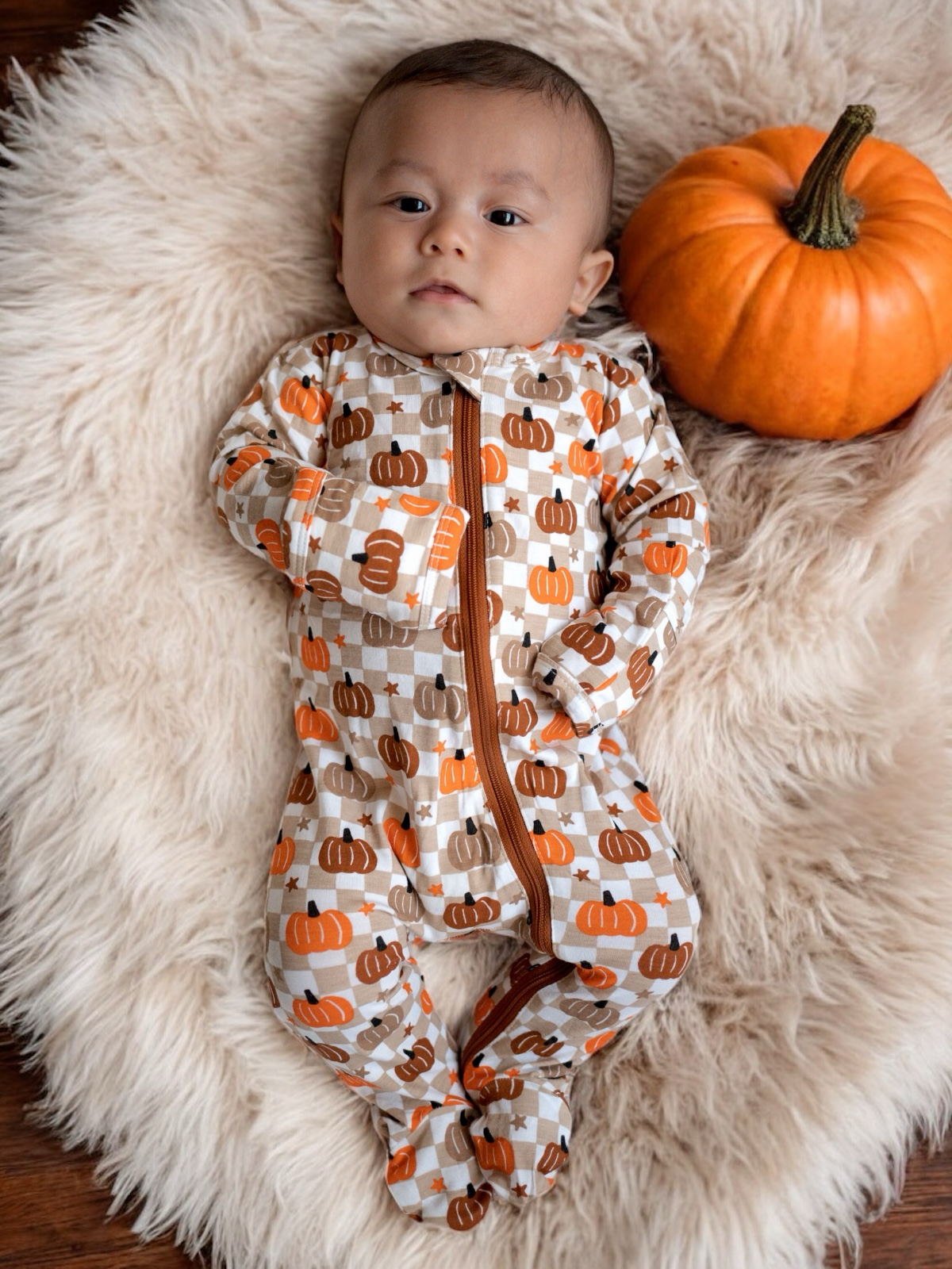 Baby in pumpkin-patterned pajamas lies on a fluffy rug next to a bright orange pumpkin.