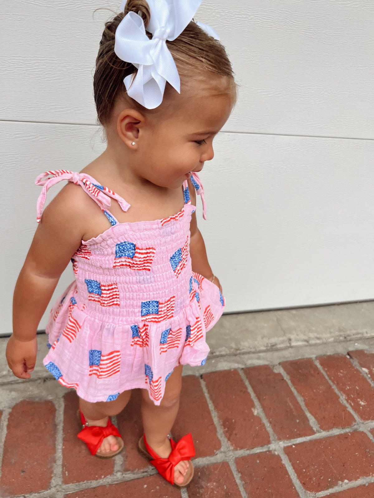 Toddler in patriotic dress with flags, white bow in hair, standing on a brick path.