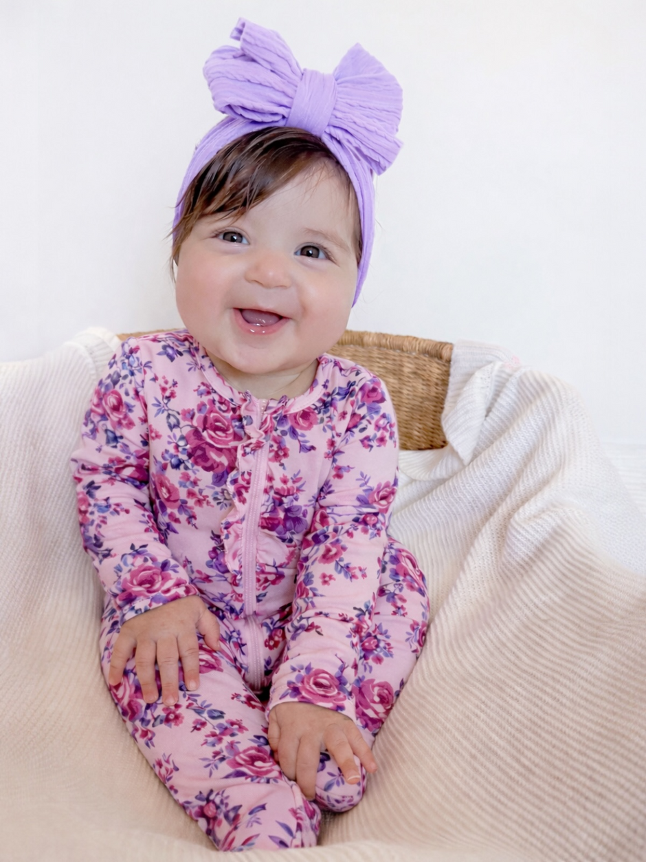 Smiling baby in floral pajamas with a purple bow headband, sitting on a cozy blanket.
