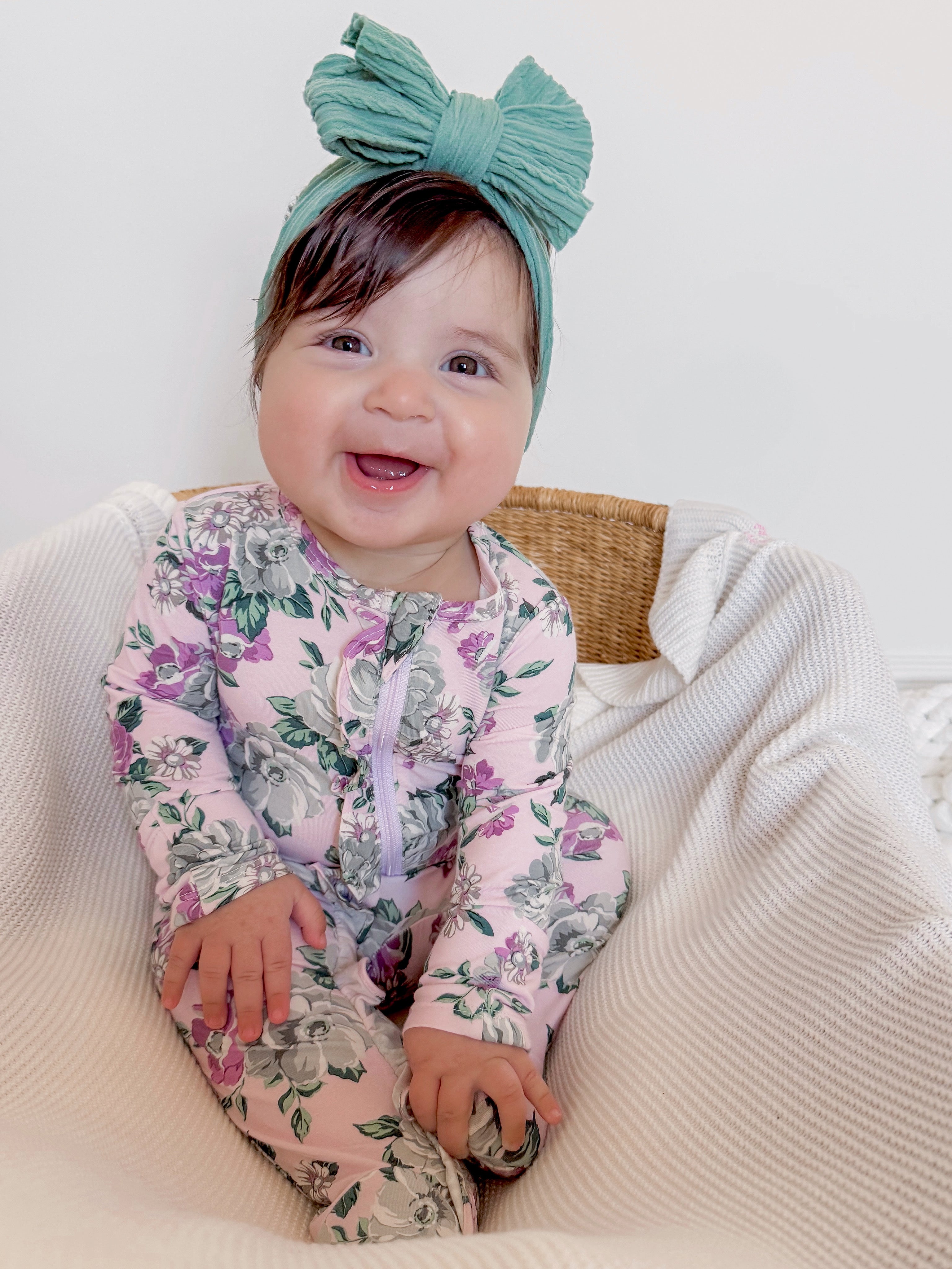 Smiling baby in floral onesie with a large teal bow, sitting on a white blanket in a wicker basket.