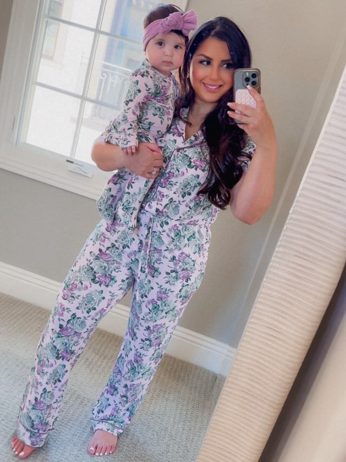 Mom and daughter in matching floral pajamas, smiling in a mirror selfie. Soft lighting and cozy setting.