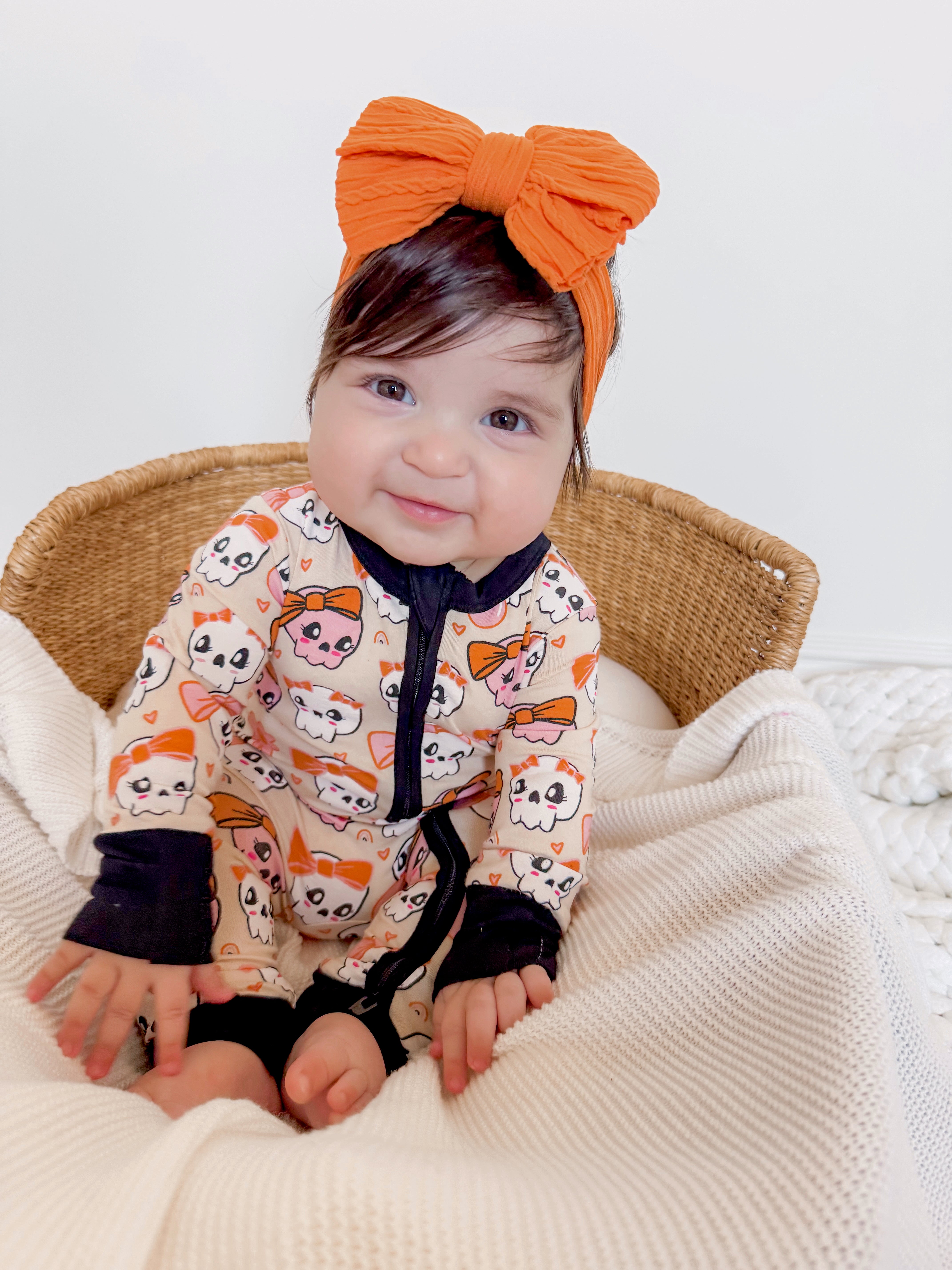 Smiling baby in colorful cat-themed onesie and large orange bow, seated on a cozy blanket in a wicker chair.