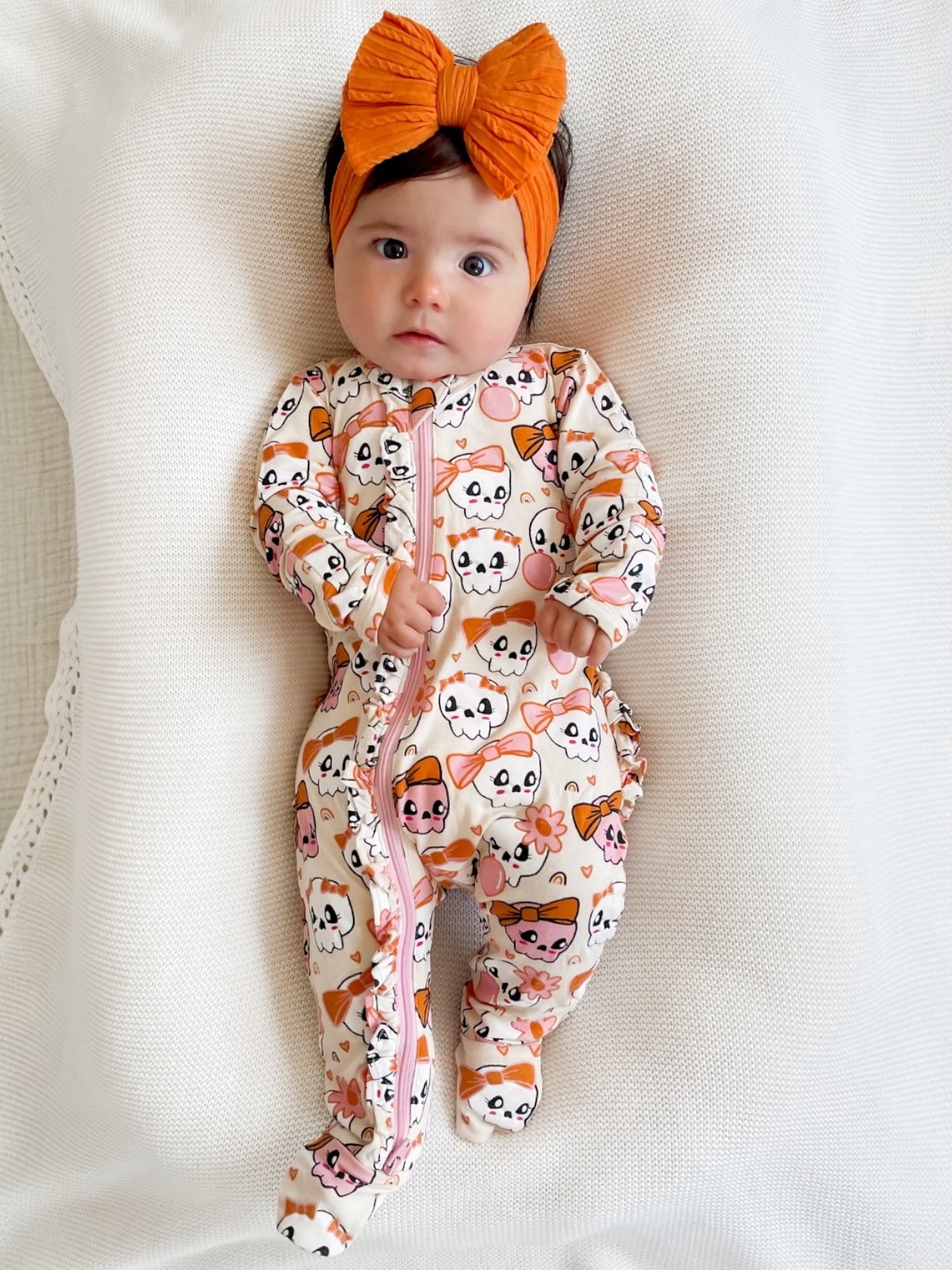 Infant in a playful skull-patterned onesie with an orange bow headband, lying on a cozy white blanket.