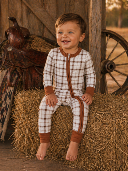 Smiling toddler in plaid pajamas sitting on a hay bale near a rustic wooden backdrop.