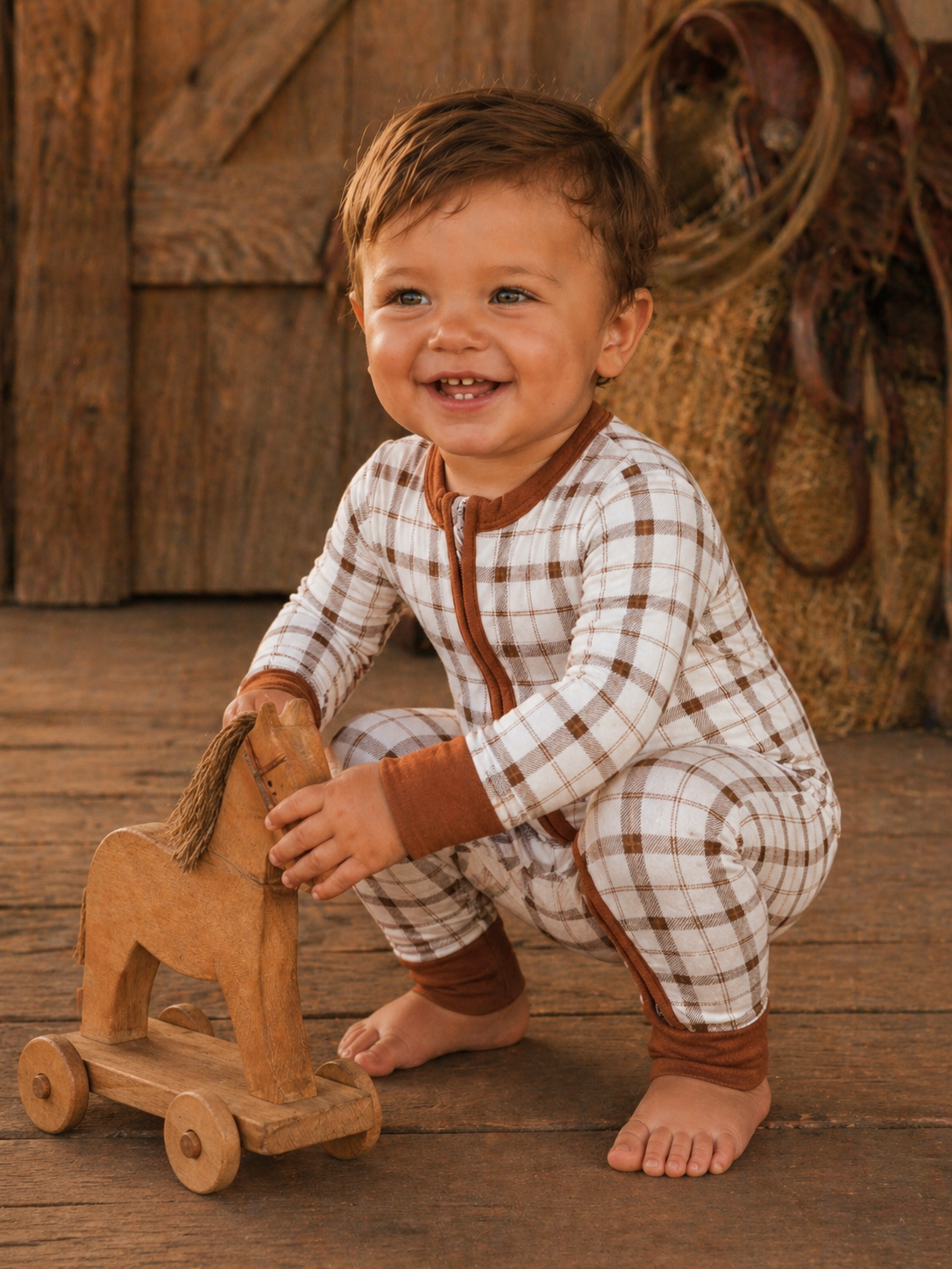Smiling toddler in plaid pajamas playing with a wooden rocking horse on rustic wooden floor.