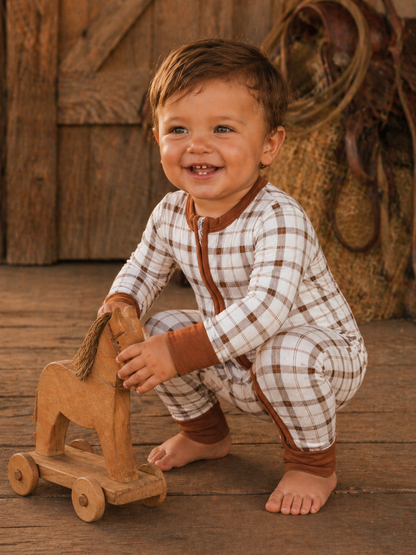 Smiling toddler in plaid pajamas playing with a wooden rocking horse on rustic wooden floor.