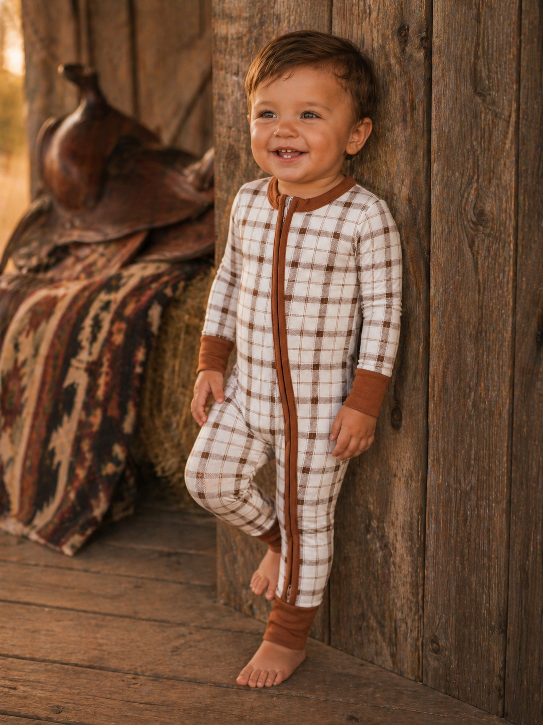 Smiling toddler in a plaid onesie, leaning against a rustic wooden wall with an earthy background.