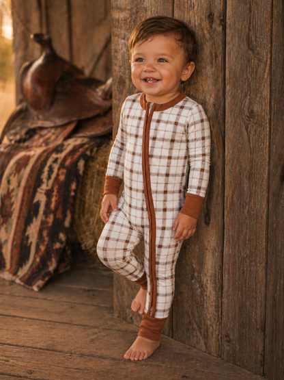 Smiling toddler in a plaid onesie, leaning against a rustic wooden wall with an earthy background.