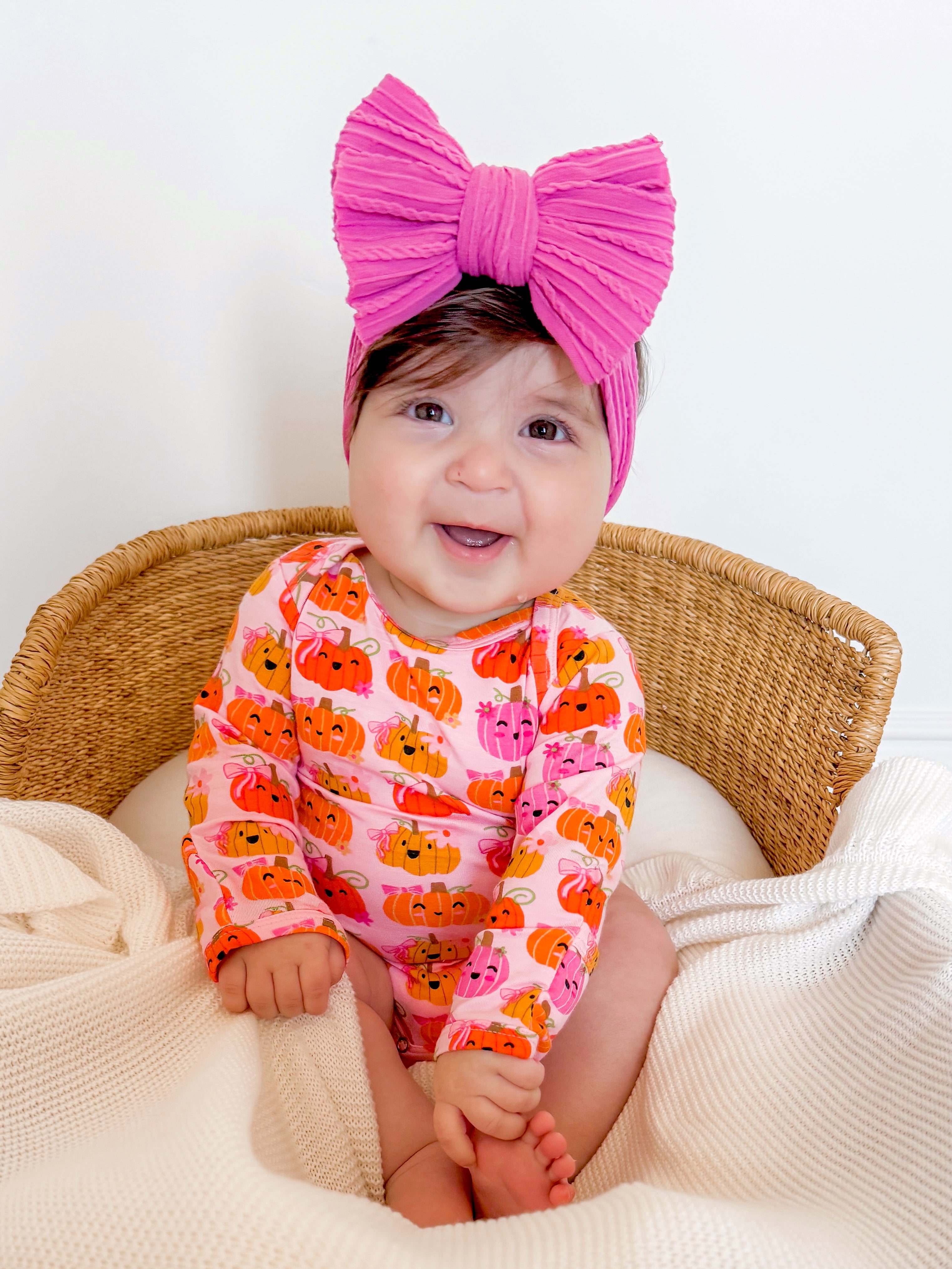 Smiling baby in a pumpkin-patterned outfit and pink bow, sitting in a woven basket with a cozy blanket.