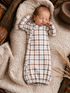 Newborn baby sleeping on a textured blanket, dressed in a brown and white plaid outfit with a hat nearby.
