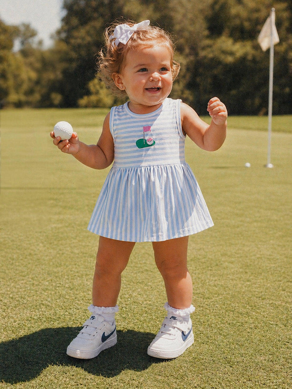 Smiling toddler in a striped dress holds a golf ball, standing on a green field with a golf flag in the background.