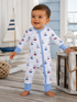 Happy toddler in a boat-patterned pajama, standing on wooden floor near toys and decor in a bright, airy room.