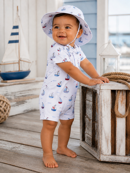 Smiling baby in a nautical-themed outfit and hat, standing beside a wooden crate and boat decor.