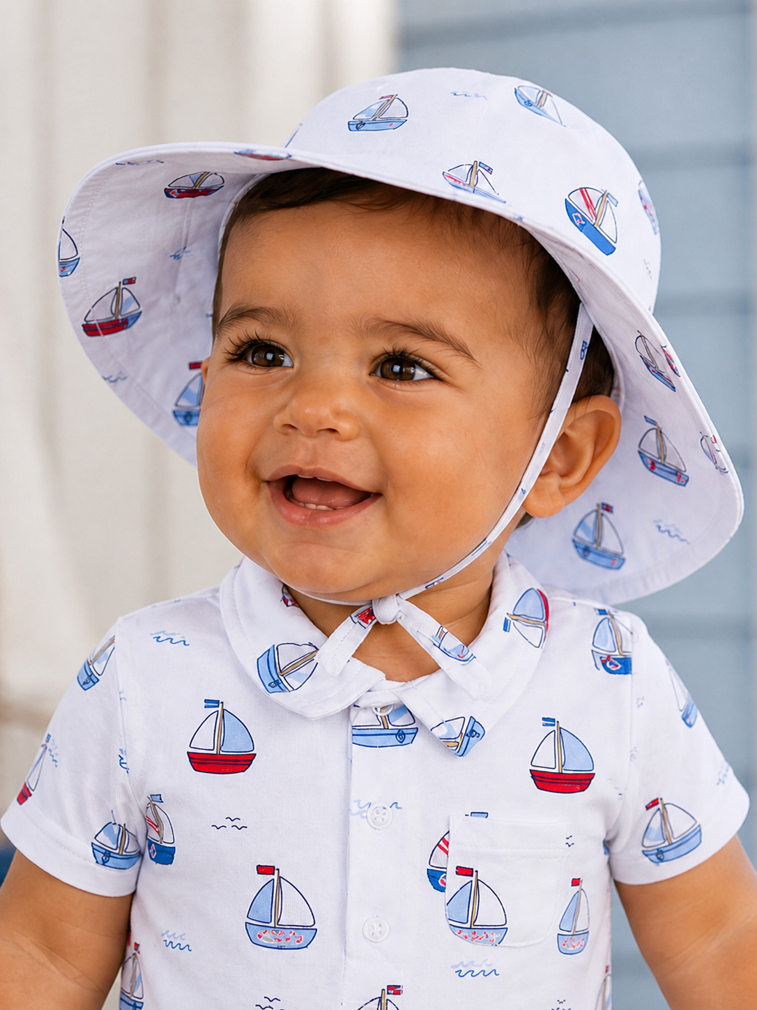 Smiling toddler in a sailboat-patterned outfit and sun hat, set against a light blue background.