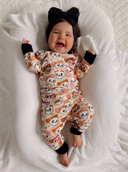 Smiling baby in cute ghost-patterned pajamas, wearing a large black bow, on a cozy blanket.