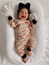 Smiling baby in cute ghost-patterned pajamas, wearing a large black bow, on a cozy blanket.
