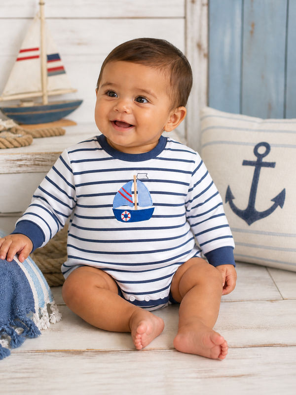 Smiling baby in a striped onesie with a sailboat design, sitting on a wooden floor next to nautical decor.