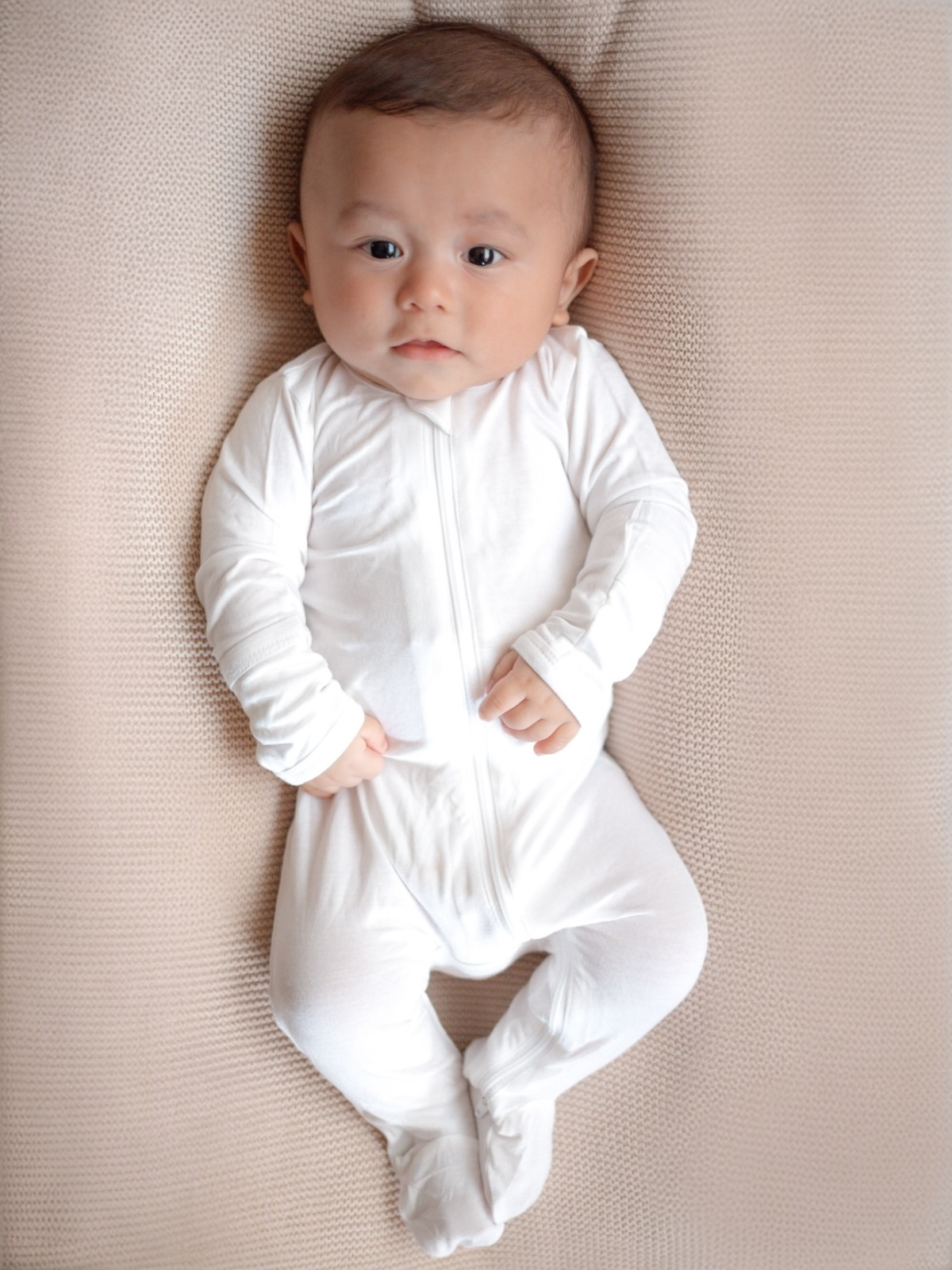 Baby in a white onesie lying on a soft, textured blanket, looking directly at the camera.