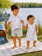 Two children walk hand in hand by a lake, wearing playful summer outfits, with a "Gone Fishing" sign nearby.