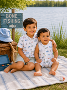 Two smiling boys sitting on a blanket by a lake, next to a "Gone Fishing" sign and a picnic basket.