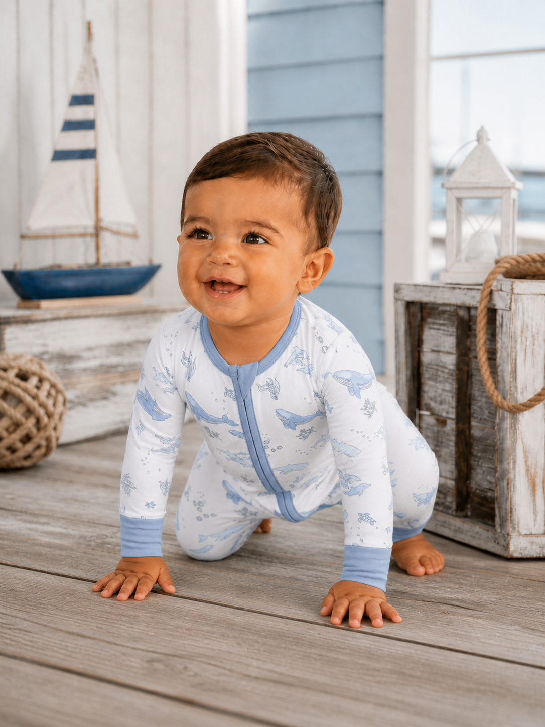 Smiling baby in whale-patterned pajamas crawling on wooden floor with nautical decor in the background.