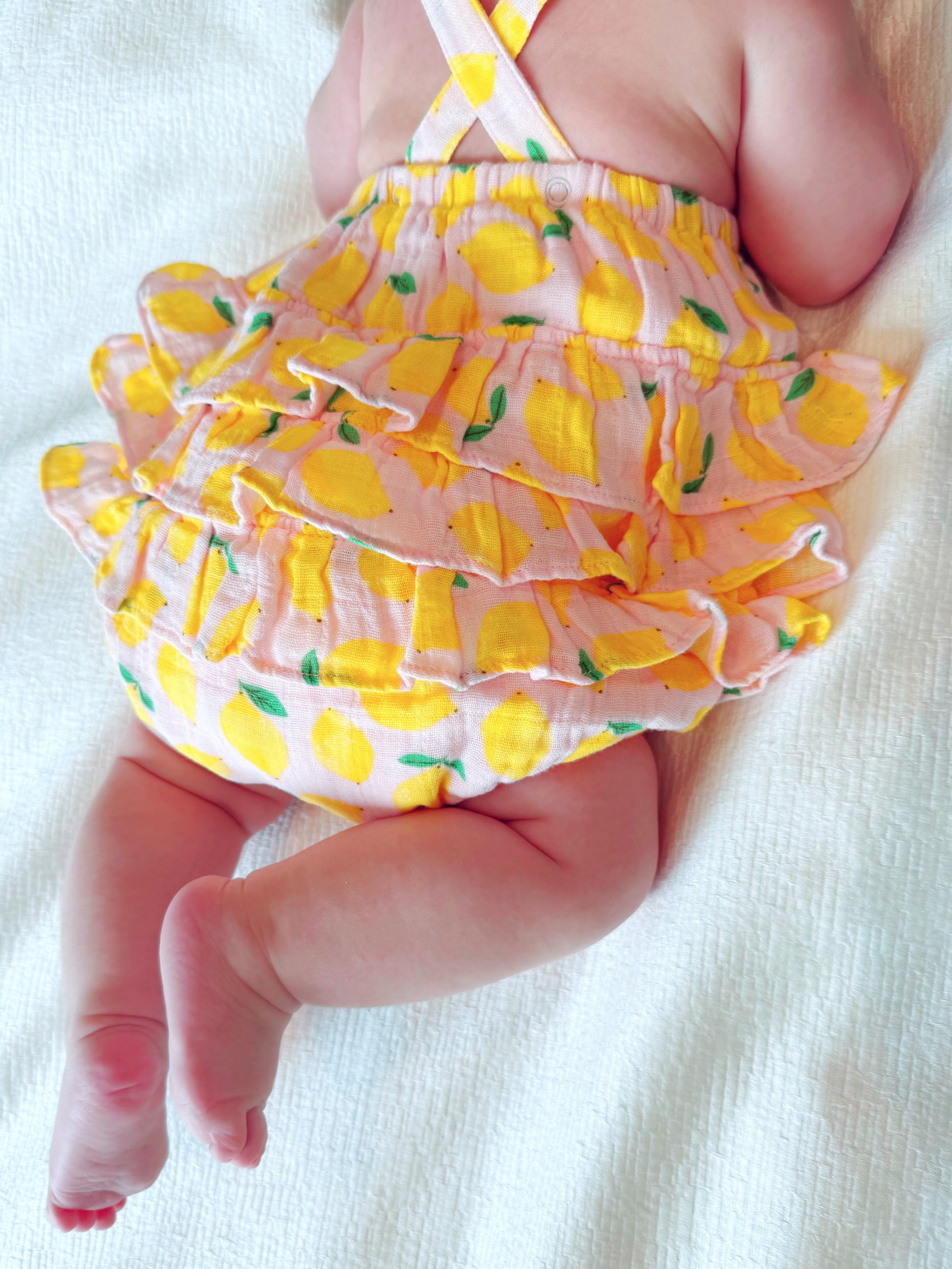 Baby wearing a lemon-patterned outfit, lying on a soft white surface, showing chubby arms and legs.