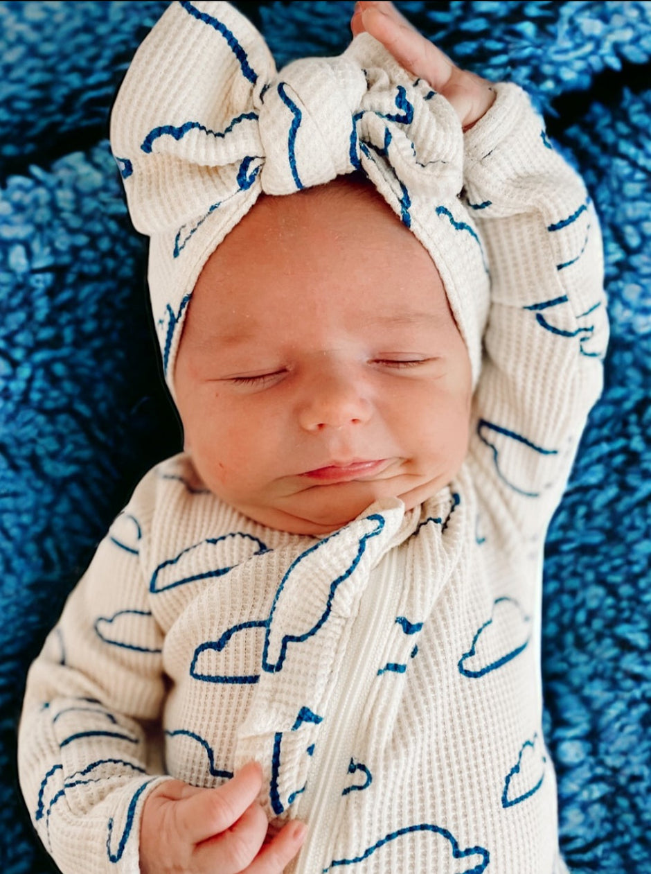 Baby asleep on blue blanket, wearing a cloud-patterned outfit and a large bow headband, with a peaceful expression.