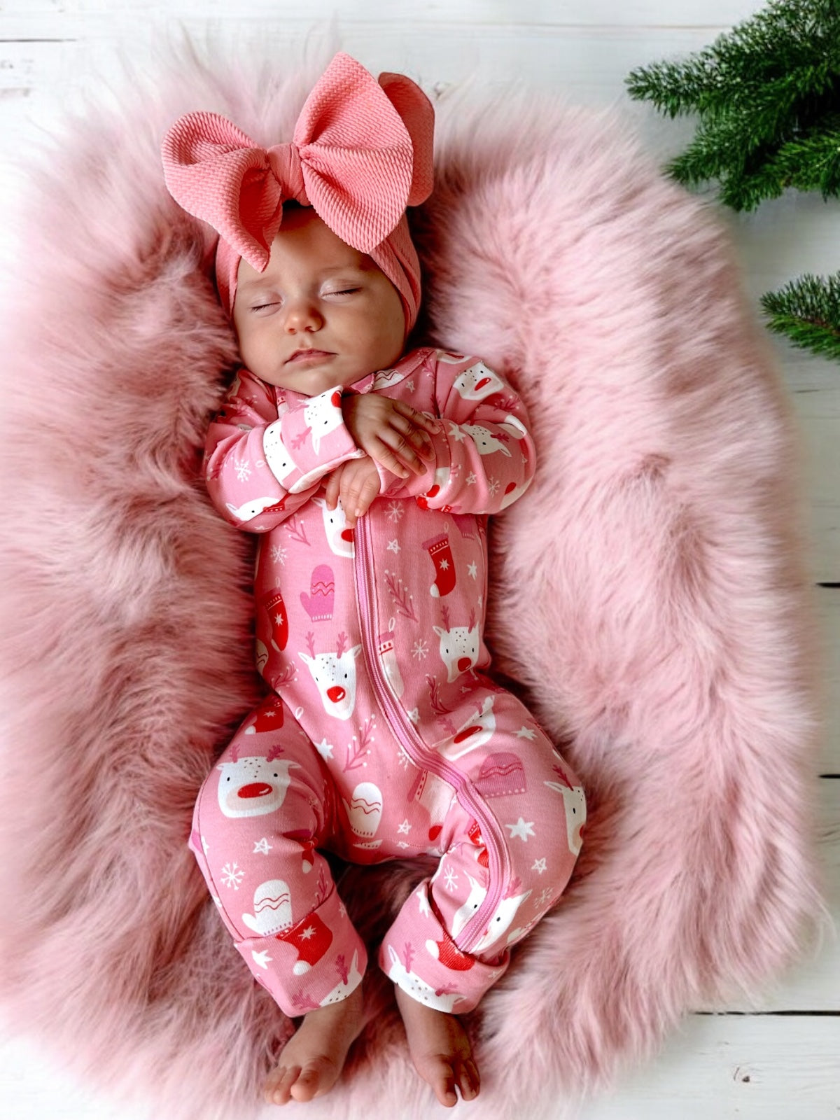 Sleeping baby in pink onesie with festive patterns, wearing a large pink bow, on a soft pink fur blanket.