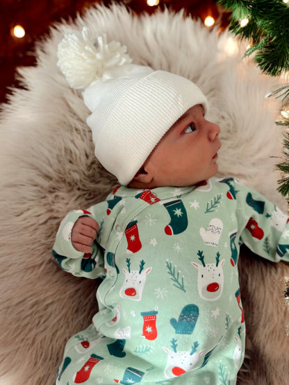 Infant in festive onesie with reindeer and mittens, wearing a white pom-pom beanie, resting on soft fur.