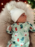 Infant in festive onesie with reindeer and mittens, wearing a white pom-pom beanie, resting on soft fur.