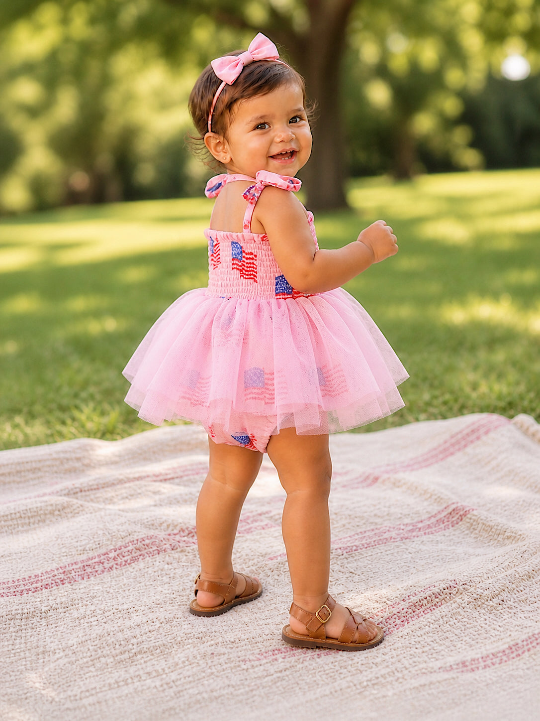 Smiling girl in a pink dress with patriotic patterns, standing on a blanket in a sunny park.