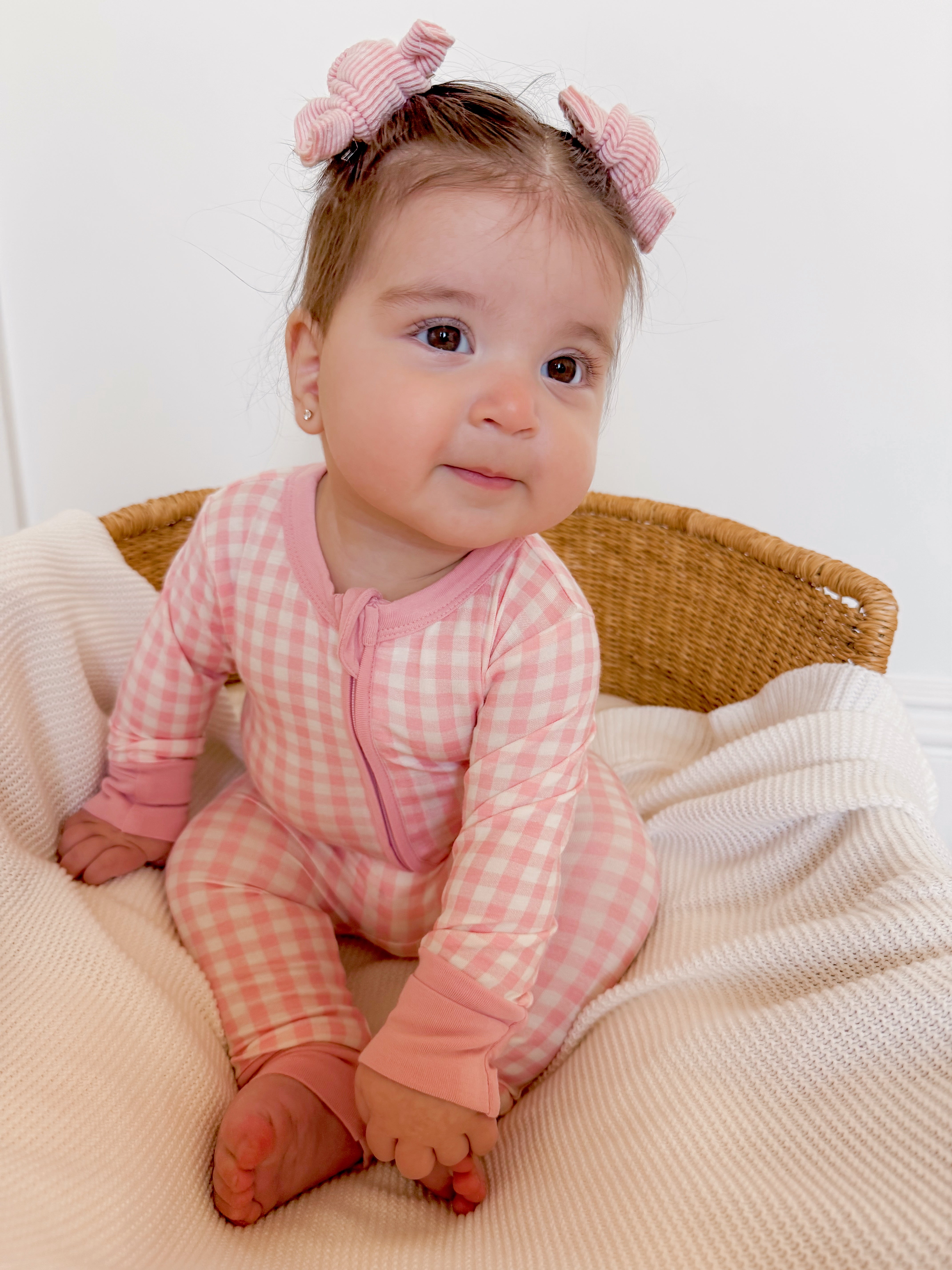 Baby girl in pink checkered pajamas with hair bows, sitting in a cozy basket on a soft blanket.