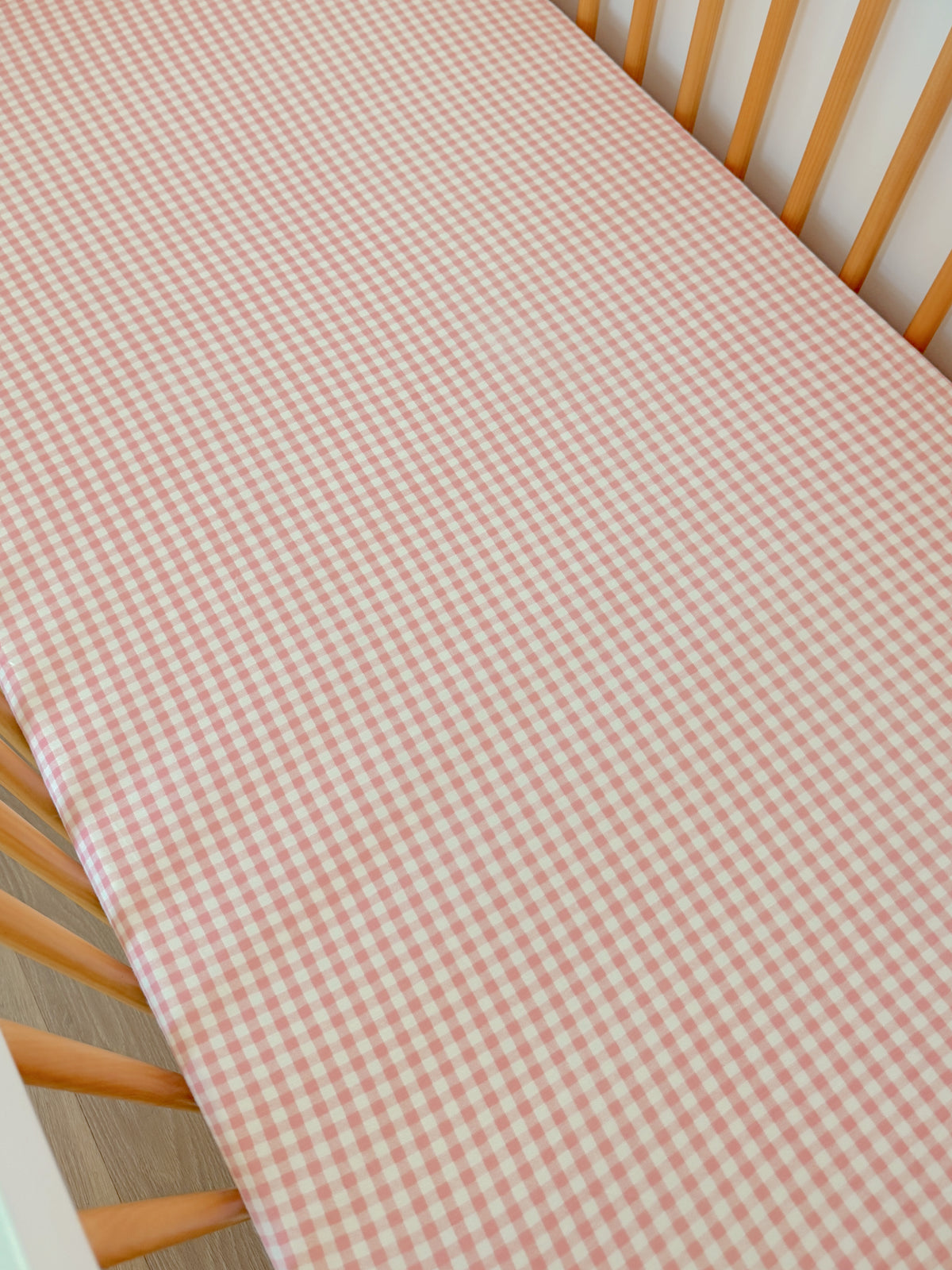 Pink and white gingham patterned crib sheet on a wooden crib.