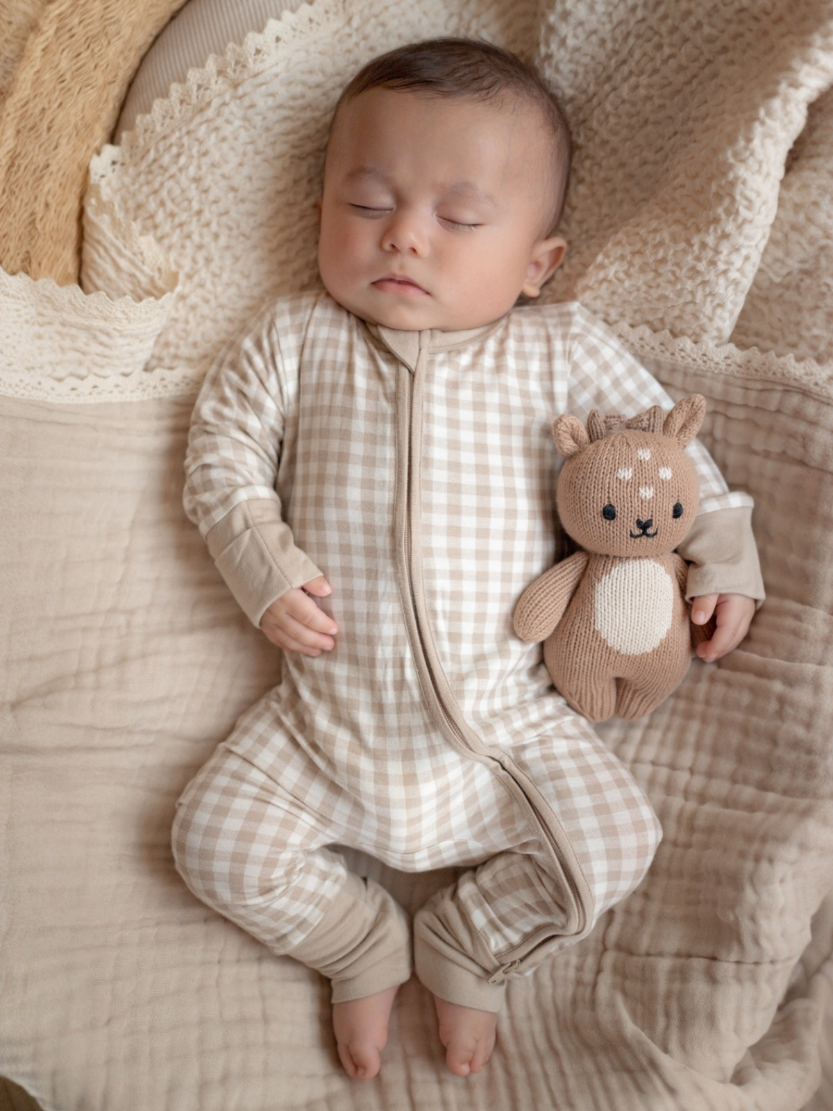 Baby in a beige plaid onesie sleeping with a knitted animal toy on a cozy blanket.