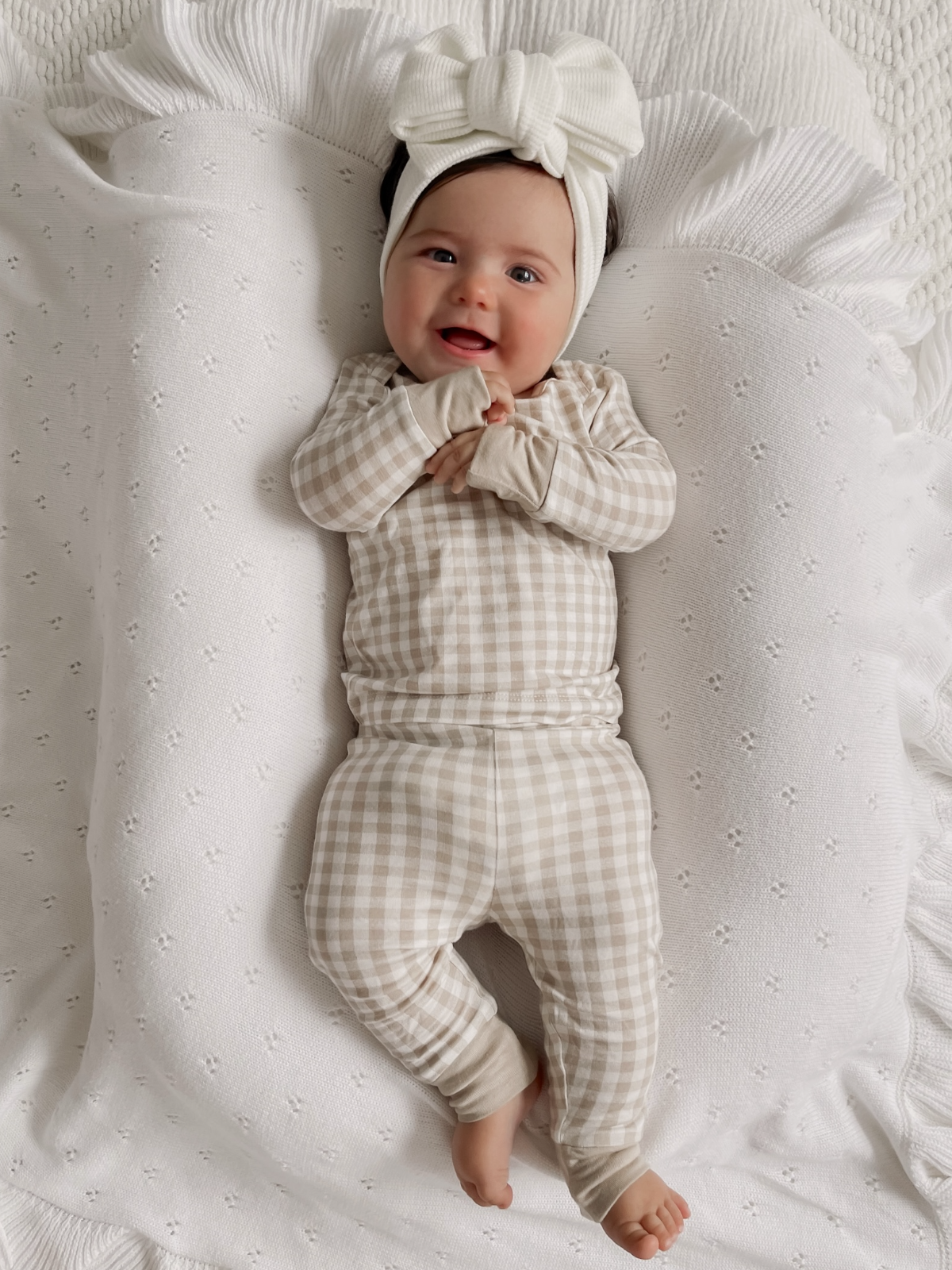 Smiling baby in a checkered outfit and bow headband, lying on a textured white blanket.