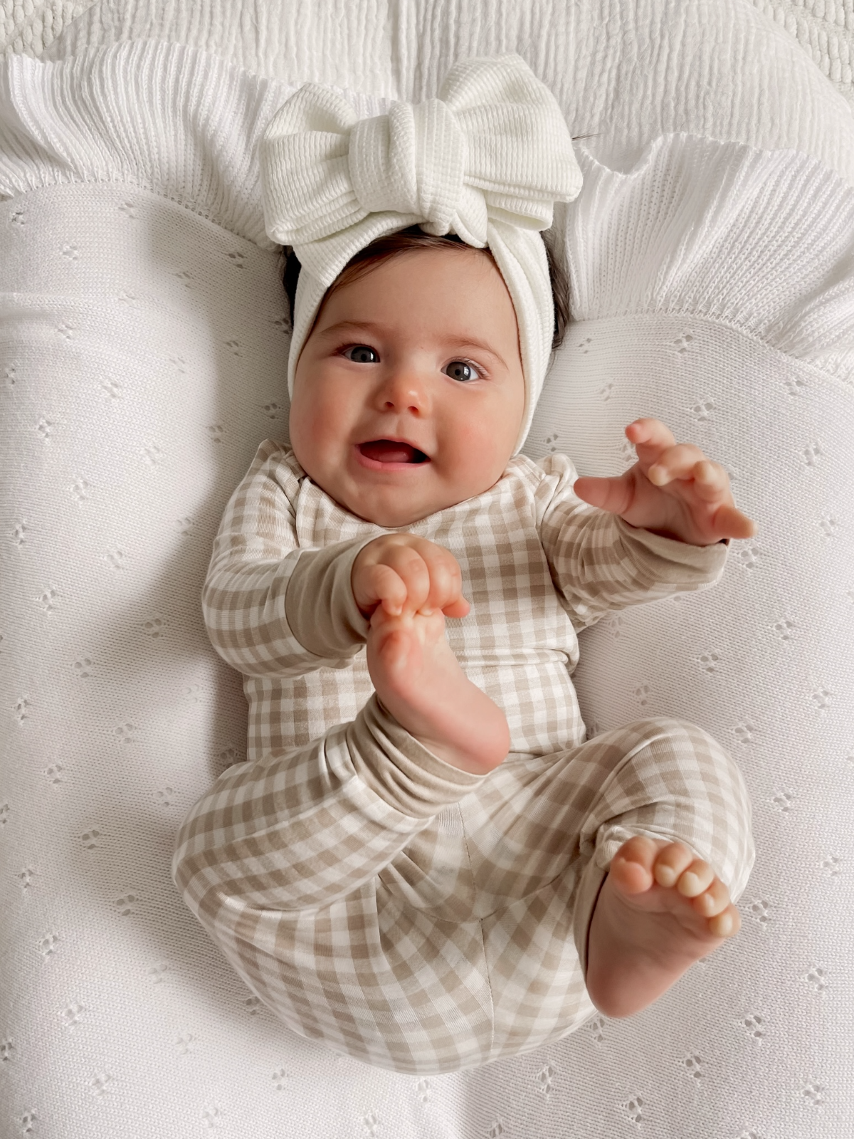 Adorable baby in a checked outfit and oversized bow, smiling while lying on a textured white blanket.