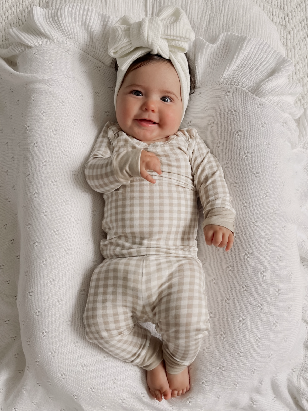 Smiling baby in a checkered outfit and large bow headband, resting on a soft white blanket.