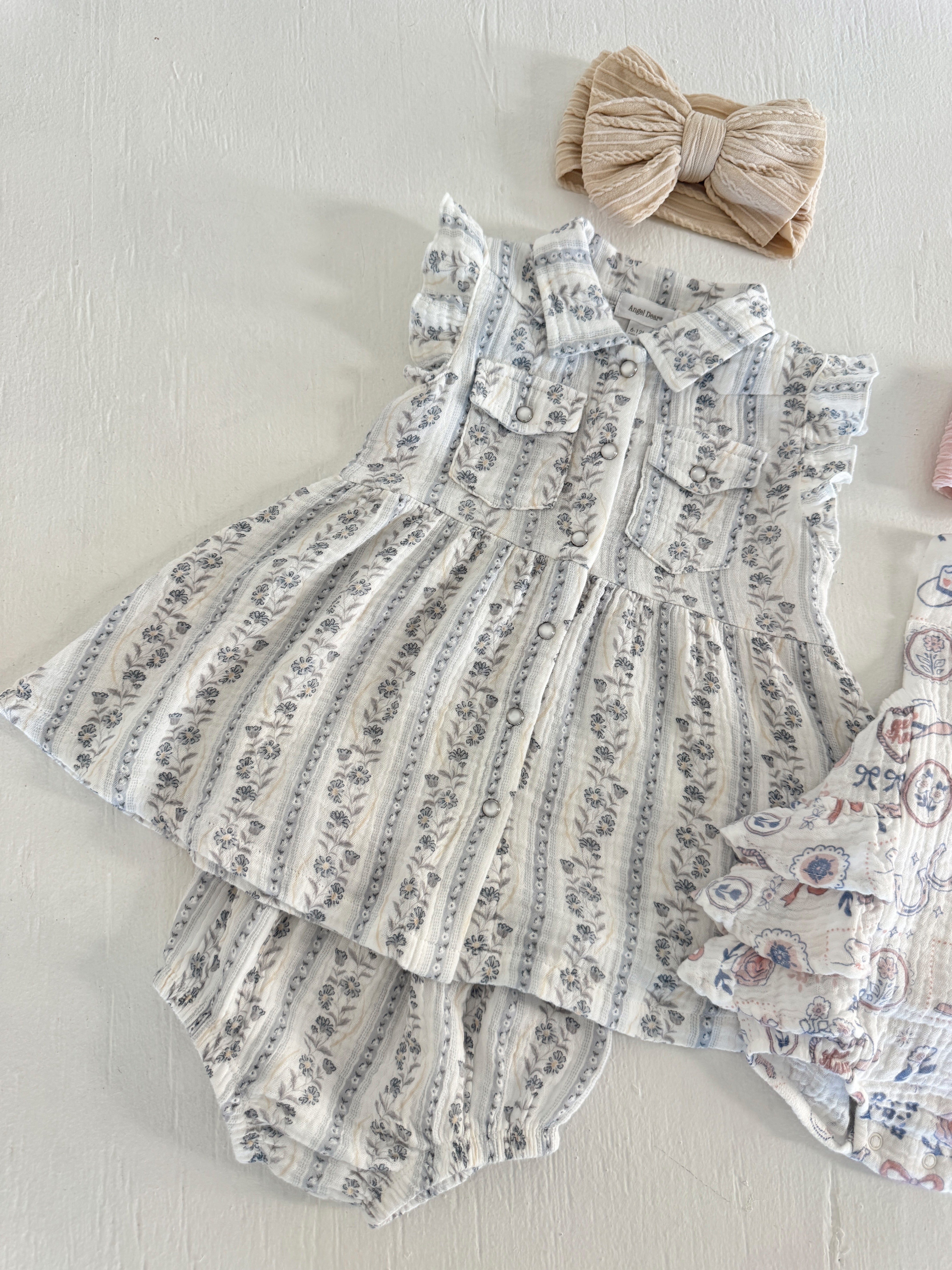 Child's sleeveless dress with ruffles, floral pattern, and two pockets, paired with a tan hair bow.