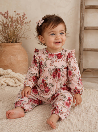 Smiling toddler in floral pink romper, sitting on a textured rug with a natural background.